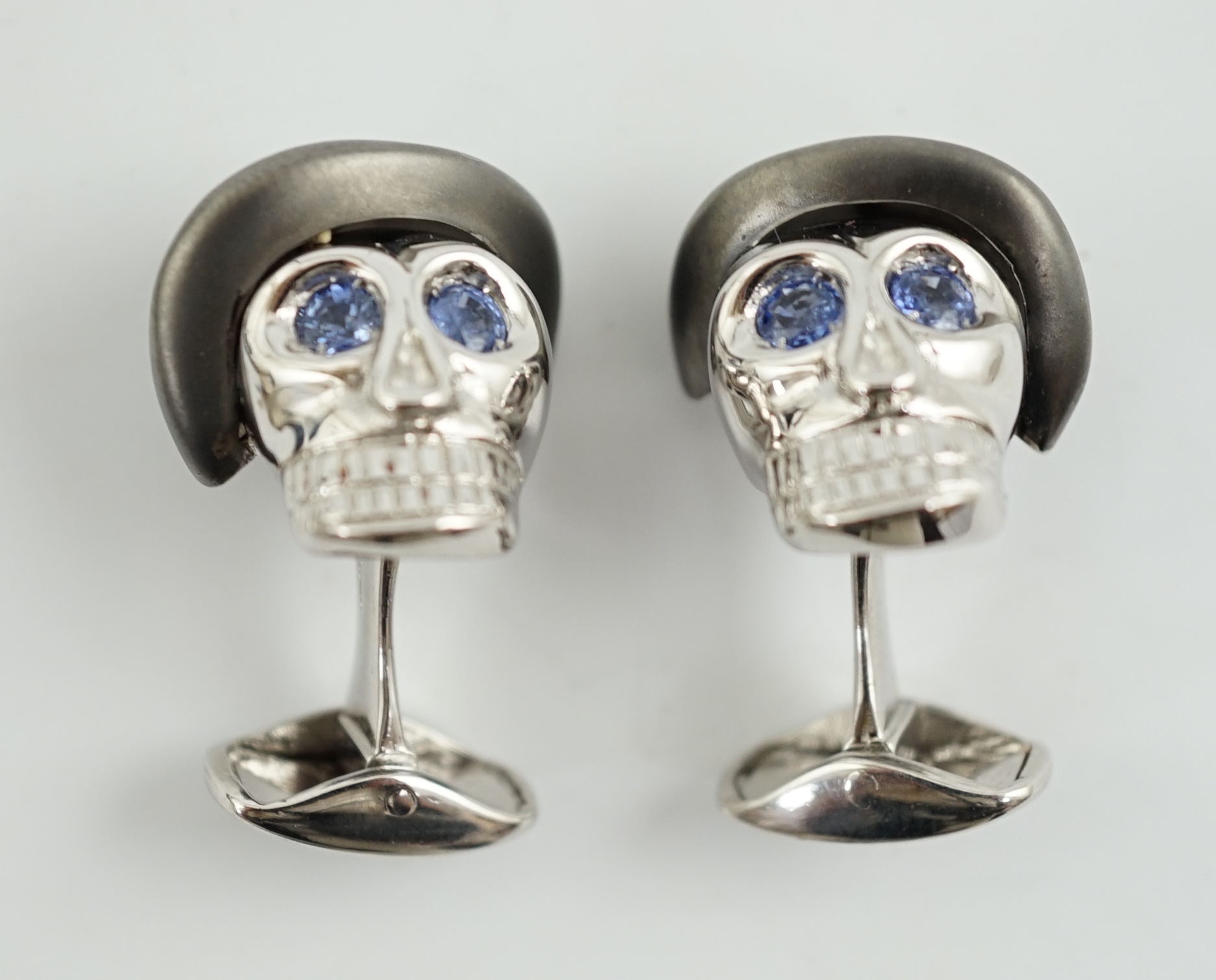 A modern pair of Longmire 18ct white gold, blackened rhodium and sapphire set novelty cufflinks, each modelled as a skull wearing a bowler hat